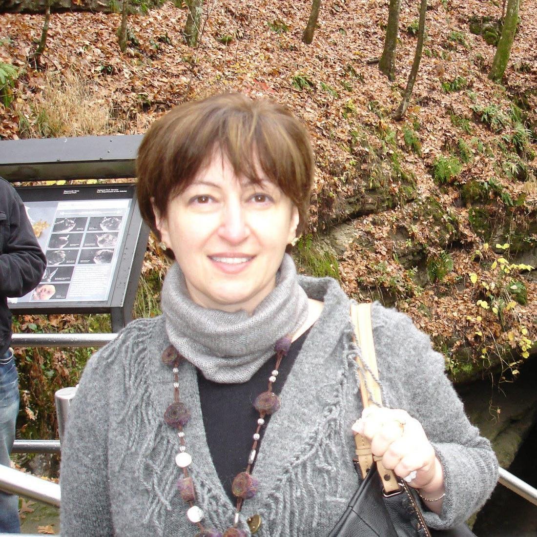 Marina Davar in a grey knit sweater and scarf, outdoors on a wooded hillside covered in autumn leaves