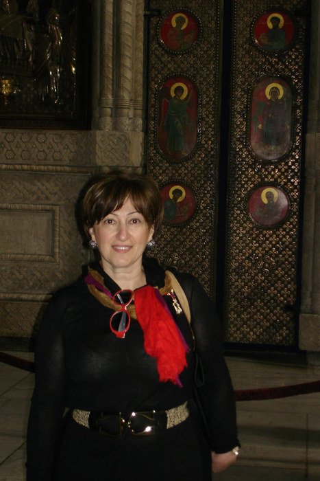 Marina Davar in a black coat and red scarf, standing in front of a set of tall doors decorated with painted icons